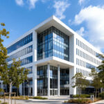 Et moderne, hvidt erhvervsbyggeri på fire etager med store glaspartier, markant udkraget tagoverhæng og en indbydende hovedindgang, omgivet af grønne træer og en solbeskinnet plads.